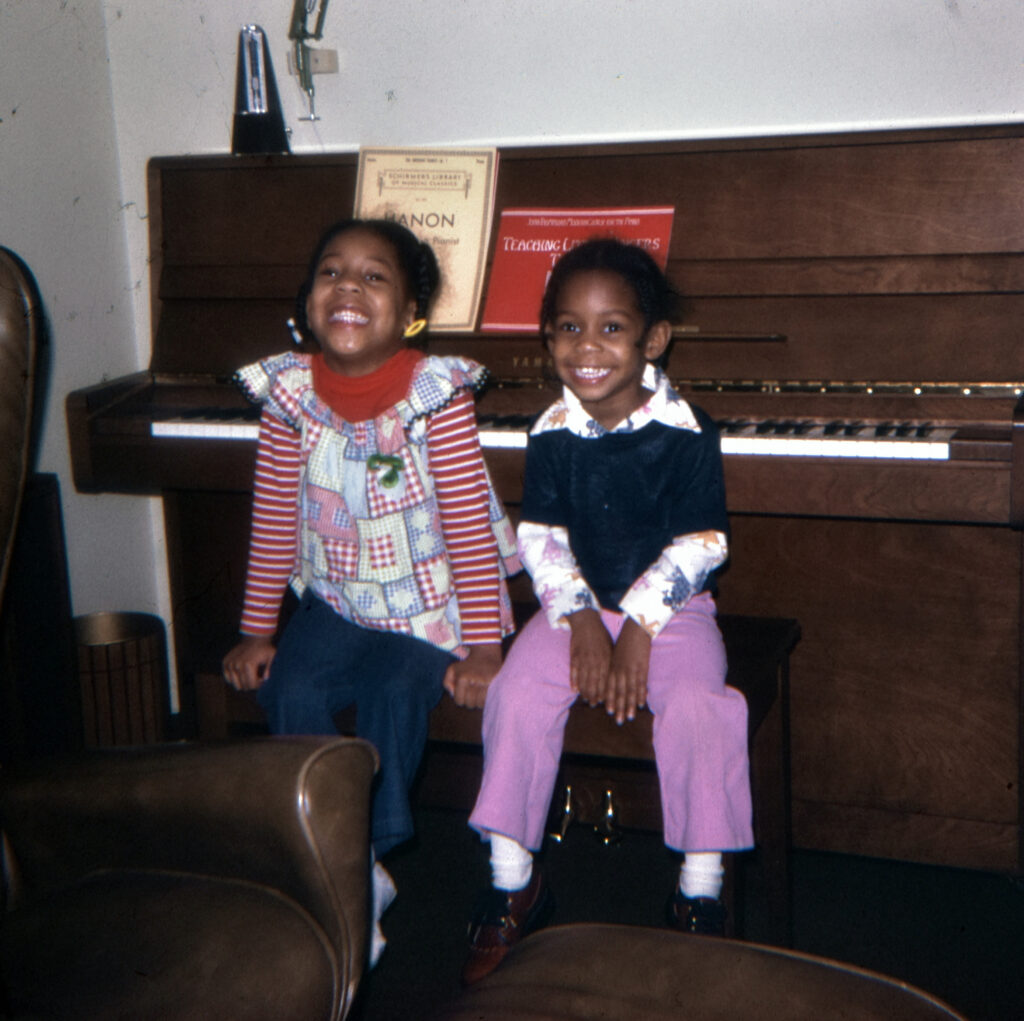 Digitized 1970s slide of two young sisters laughing on a piano bench in front of an upright piano with a metronome and lesson books